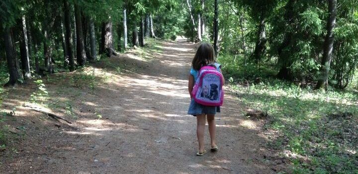 Tyttö kävelee metsäpolkua reppu selässä. Kuva otettu takaapäin.