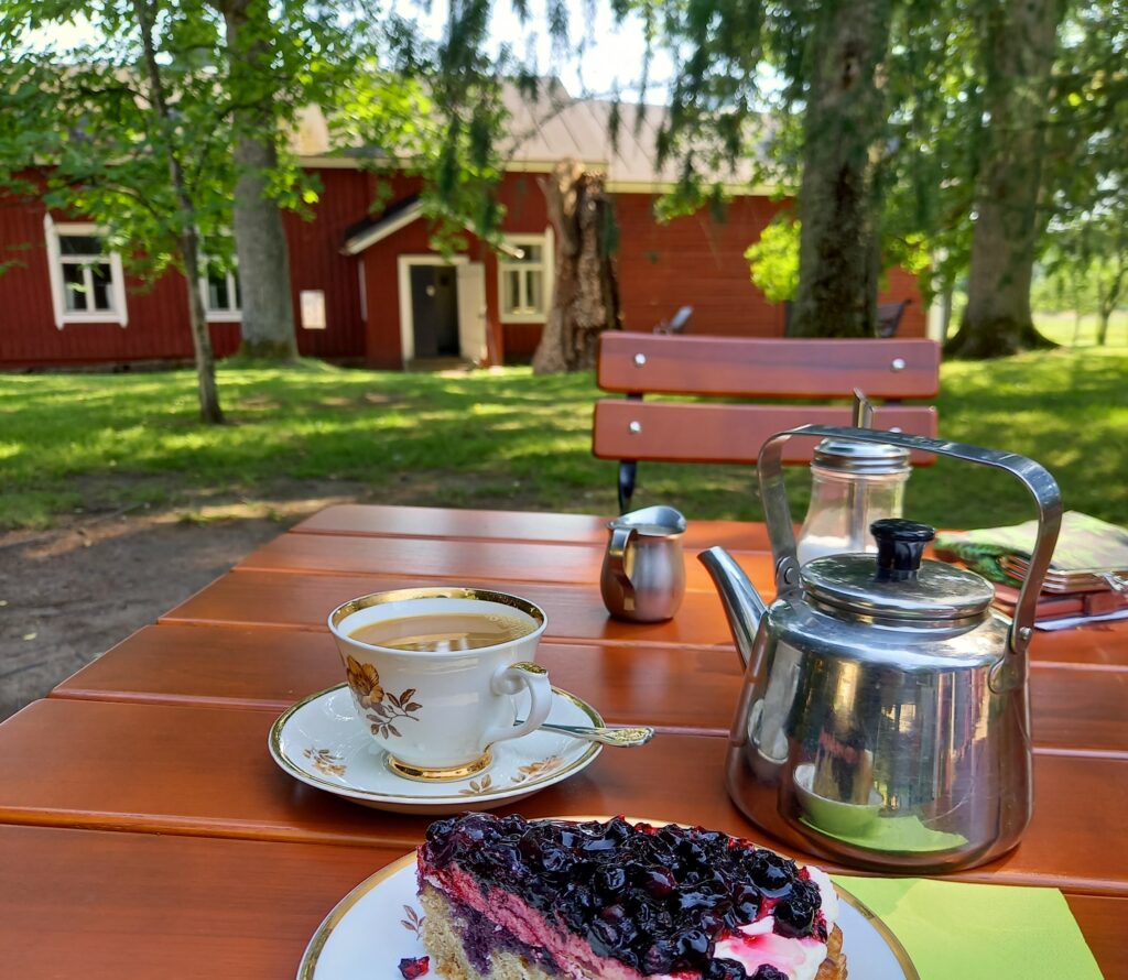 Pihapöydälle on katettu Myrna-kuppi, kahvipannu ja kakkuviipale. Tautalla on vihreä piha ja punamullalla maalattu talo.
