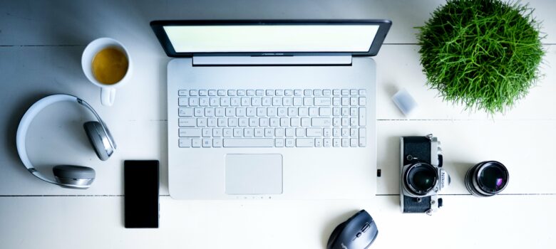 Kannettava tietokone, hiiri, kamera, kännykkä ja kahvikuppi valkoisella pöydällä.