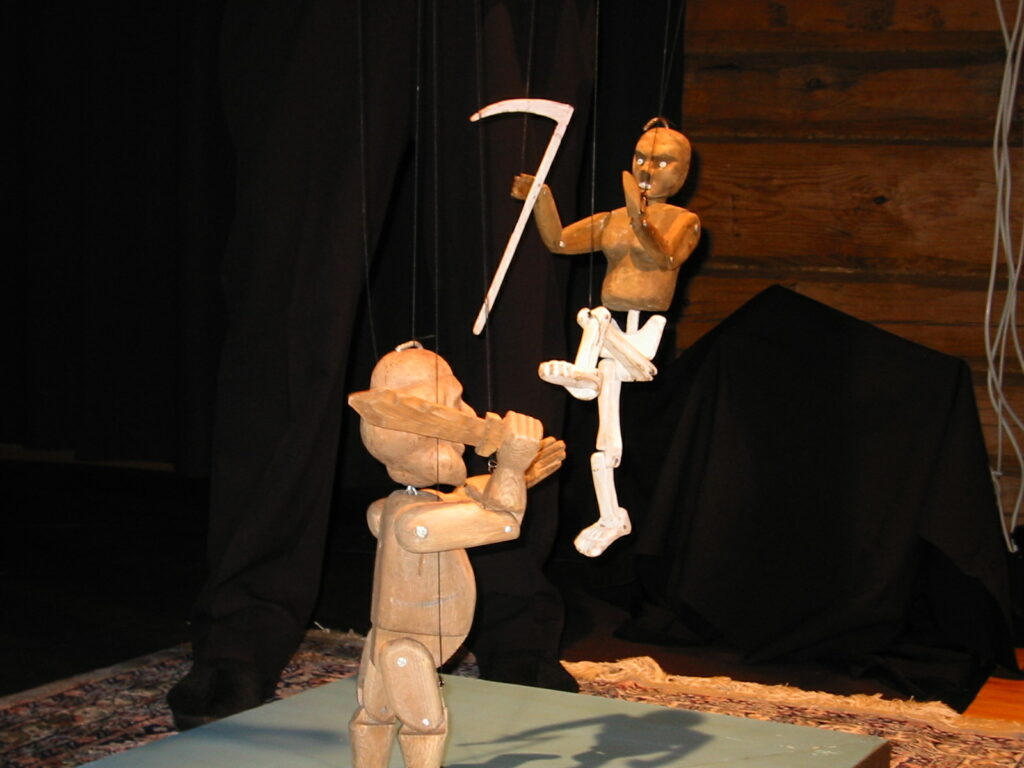 Kuvassa kaksi marionettia käyvät kamppailuun. Toisella on kädessään miekka ja toisella viikate.