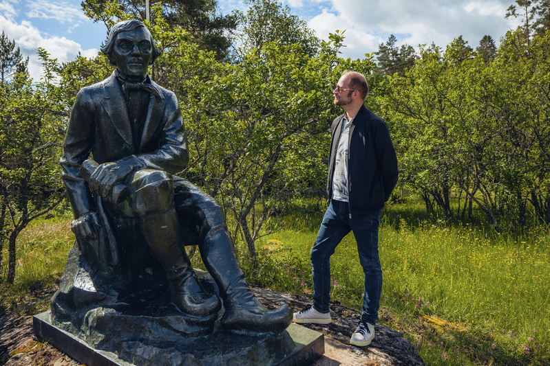 Kuvassa on Elias Lönnrotin prossinen patsas Paikkarin torpalta. Tumma patsas istuu kalliolla matalien kriikunapuiden keskellä. Mies aurinkolaseissa ihailee patsasta.