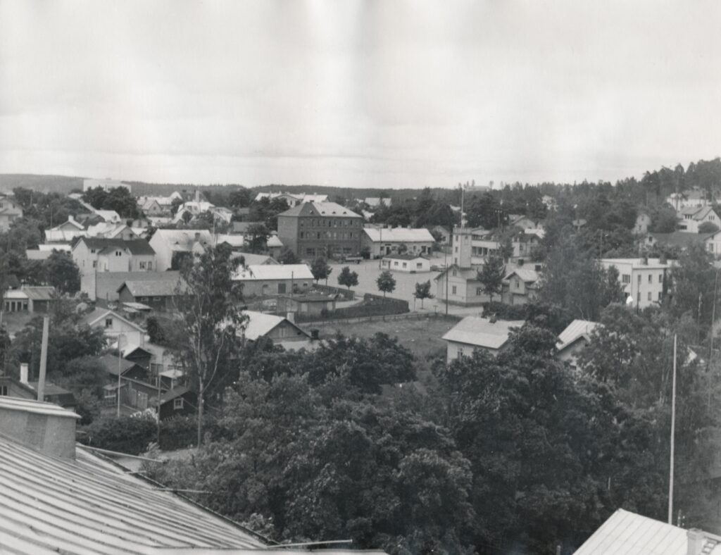 Mustavalkoisessa kuvassa on Lohjan keskustaa. Kuva on otettu talon katolta nykyisen Prisman kauppakeskuksen suunnalta. Kuvan keskellä on tori ja sen laidalla Hilnerin kauppatalo.