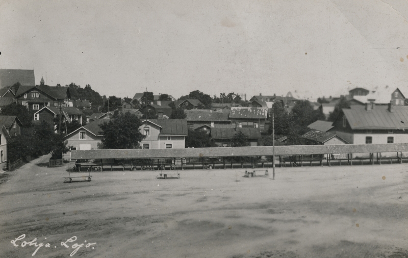 Mustavalkoisessa kuvassa on etualalla tyhjä hiekkakenttä, Lohjan tori, jonka laidalla on pitkä katettu rivi tyhjiä myyntikojuja. Taustalla on Lohjan keskustan puutaloja.