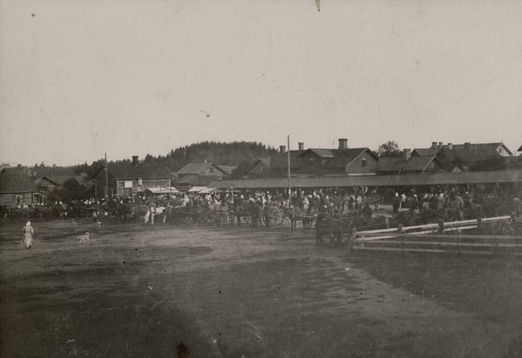 Mustavalkoisessa kuvassa on suuri joukko ihmisiä ja hevosia vaunuineen aukiolla. Taustalla on puutaloja.