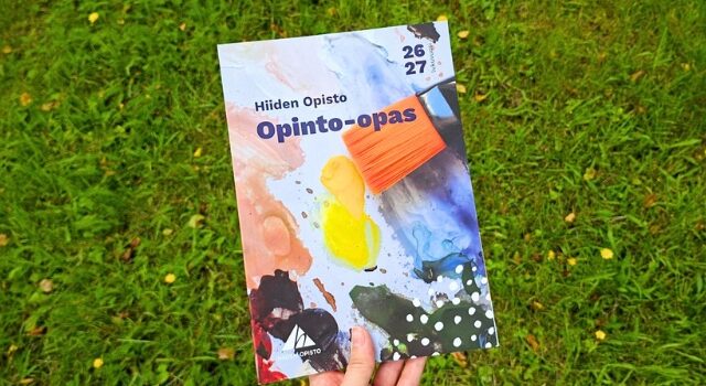Käsi pitelee Hiiden opiston opinto-opasta. Taustalla nurmikko ja kukkia.