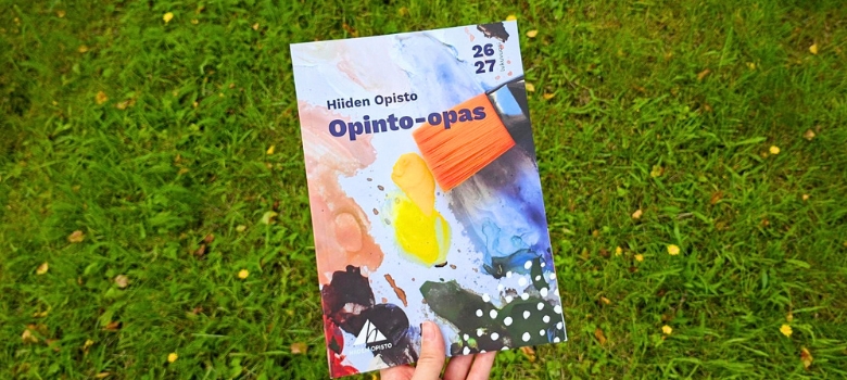 Käsi pitelee Hiiden opiston opinto-opasta. Taustalla nurmikko ja kukkia.